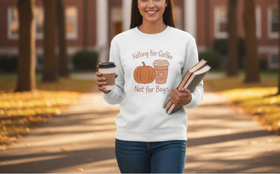 Person wearing a white sweatshirt with a pumpkin and coffee cup design, holding a coffee cup and books outdoors.